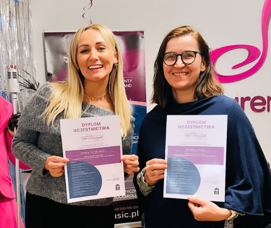 Dwie nauczycielki śpiewu z Empire Music School z dyplomami uczestnictwa po zakończonym światowym szkoleniu wokalnym w Gliwicach, organizowanym przez Stowarzyszenie Wokalistów Mix. Uśmiechnięte, stoją na tle logo szkoły i plakatu wydarzenia.